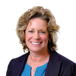A woman with curly blonde hair smiles, wearing a blue blouse and dark blazer against a plain background.